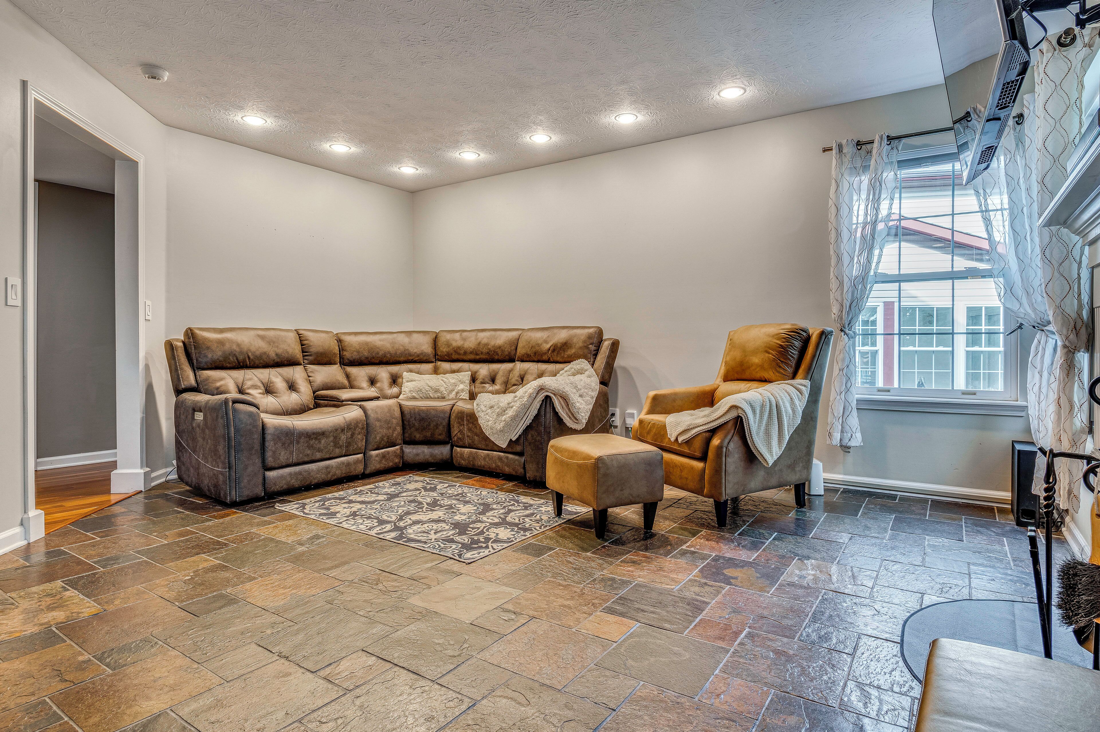 A corner living room with a sectional sofa, a single armchair, a patterned rug, and a large window with curtains.