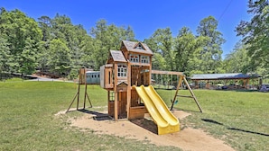 A wooden playhouse with a yellow slide in a grassy park area. - Screened Porch + Home Office: Modern Ellijay Cabin (Ellijay)