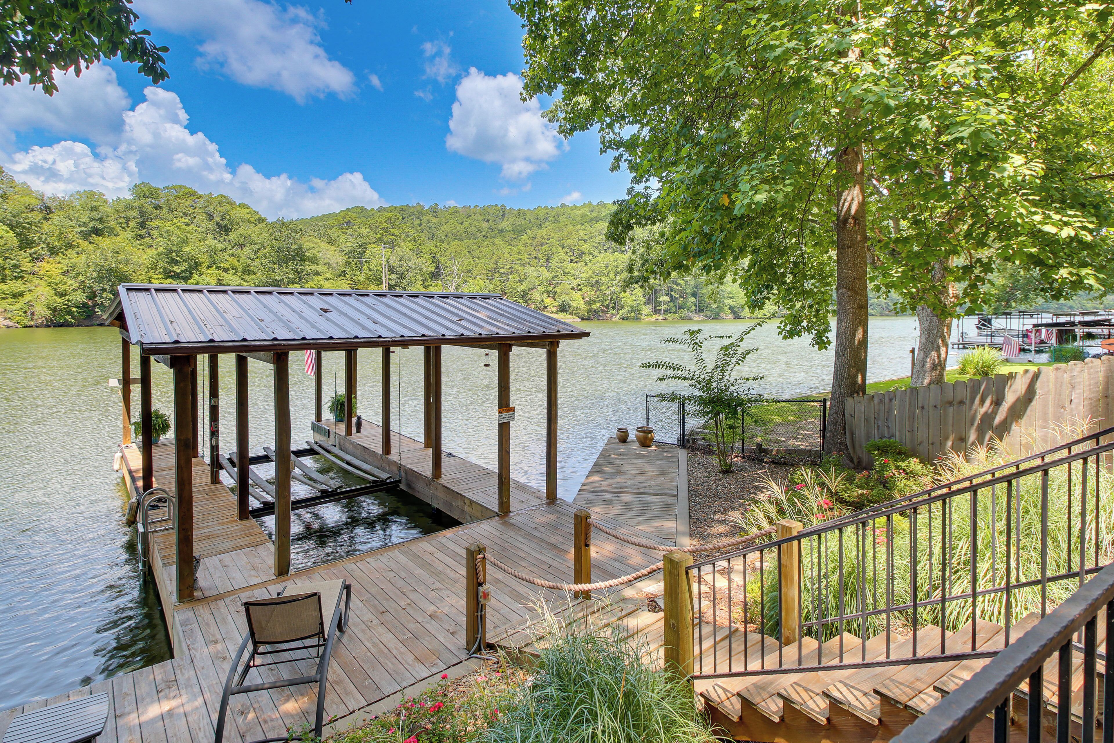 A wooden dock with a covered seating area, a small pond, and a view of a lake and forested hills.