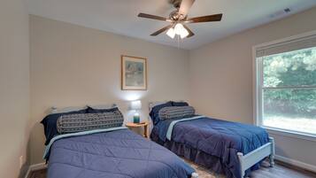 A bedroom with two beds, a ceiling fan, a nightstand with a lamp, and a window with a view of trees.