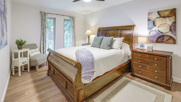Quarto com cama de madeira, mesa de cabeceira, cômoda, ventilador de teto, janela com cortinas e um quadro na parede.