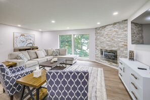Ein modernes Wohnzimmer mit einem Ecksofa, einem Couchtisch, gemusterten Sesseln, einem Kamin und großen Fenstern.