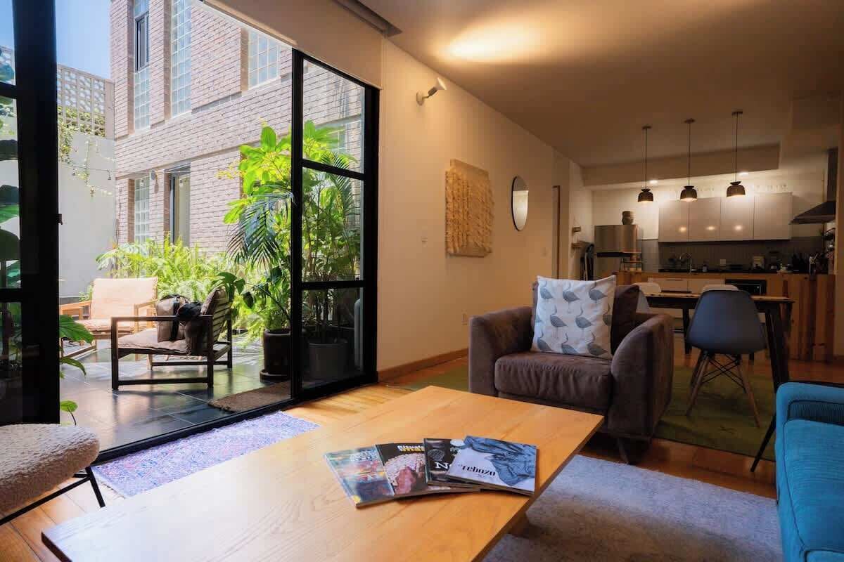 A modern living room with a large sliding glass door leading to a balcony, a dining area, and a kitchen with pendant lighting.