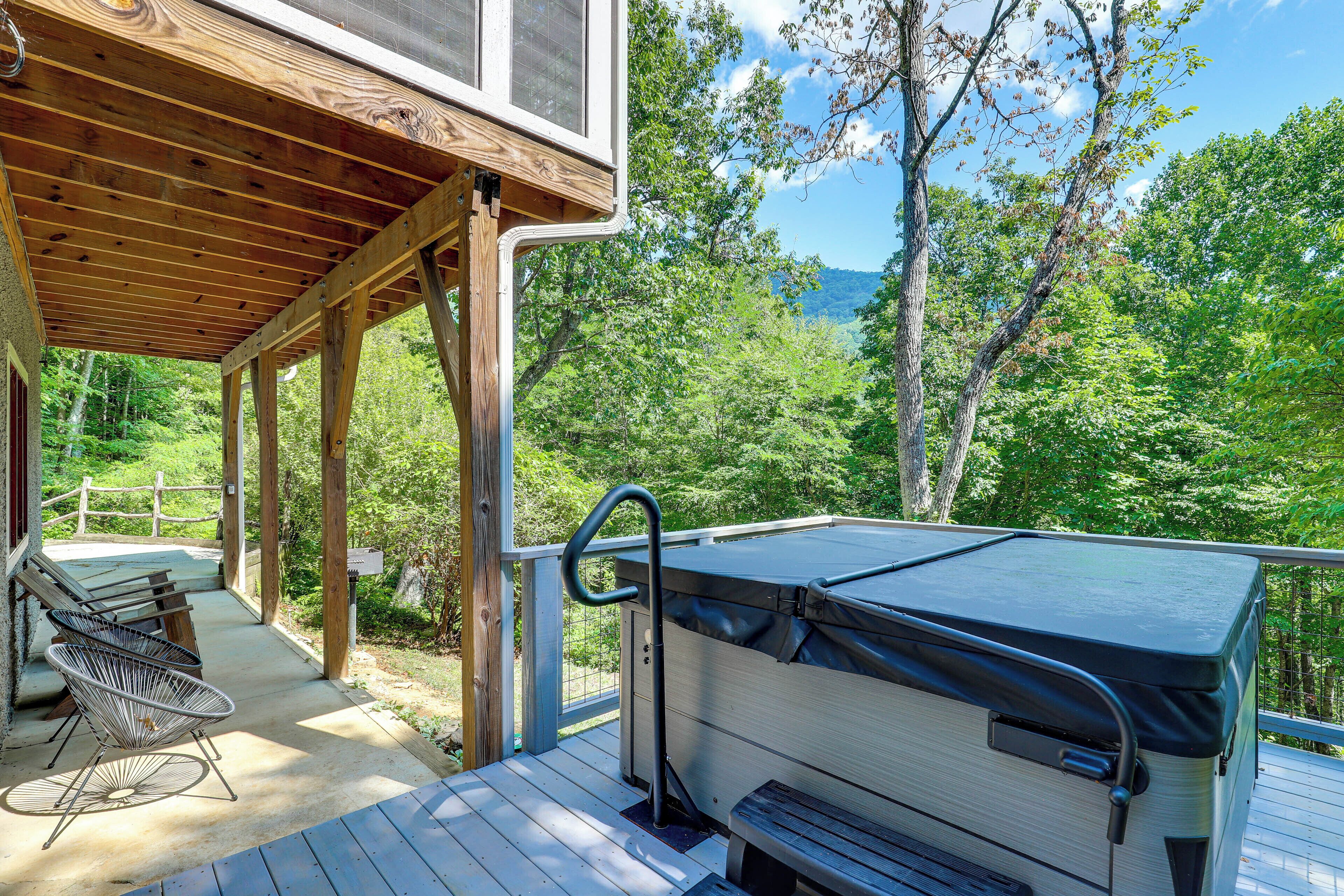 A covered deck with a hot tub, outdoor seating, and a view of a forested area.