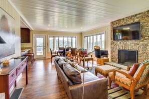 A spacious living room with a stone fireplace, a television mounted on the wall, and a large window offering a view of the outdoors. - Mountain Home Near Lake Glenville w/ Scenic Views (Cullowhee)