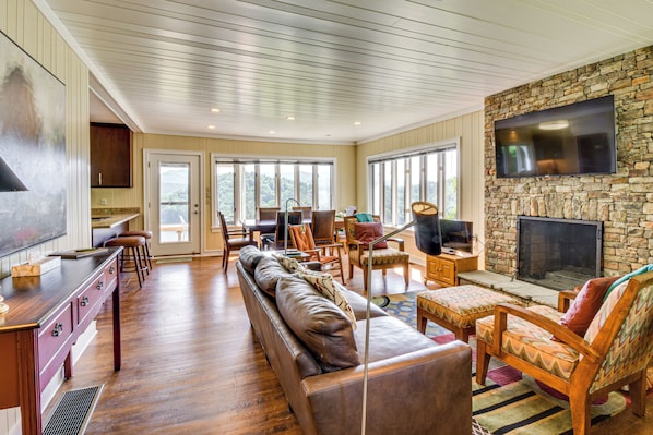 A spacious living room with a stone fireplace, a television mounted on the wall, and a large window offering a view of the outdoors. - Mountain Home Near Lake Glenville w/ Scenic Views (Cullowhee)