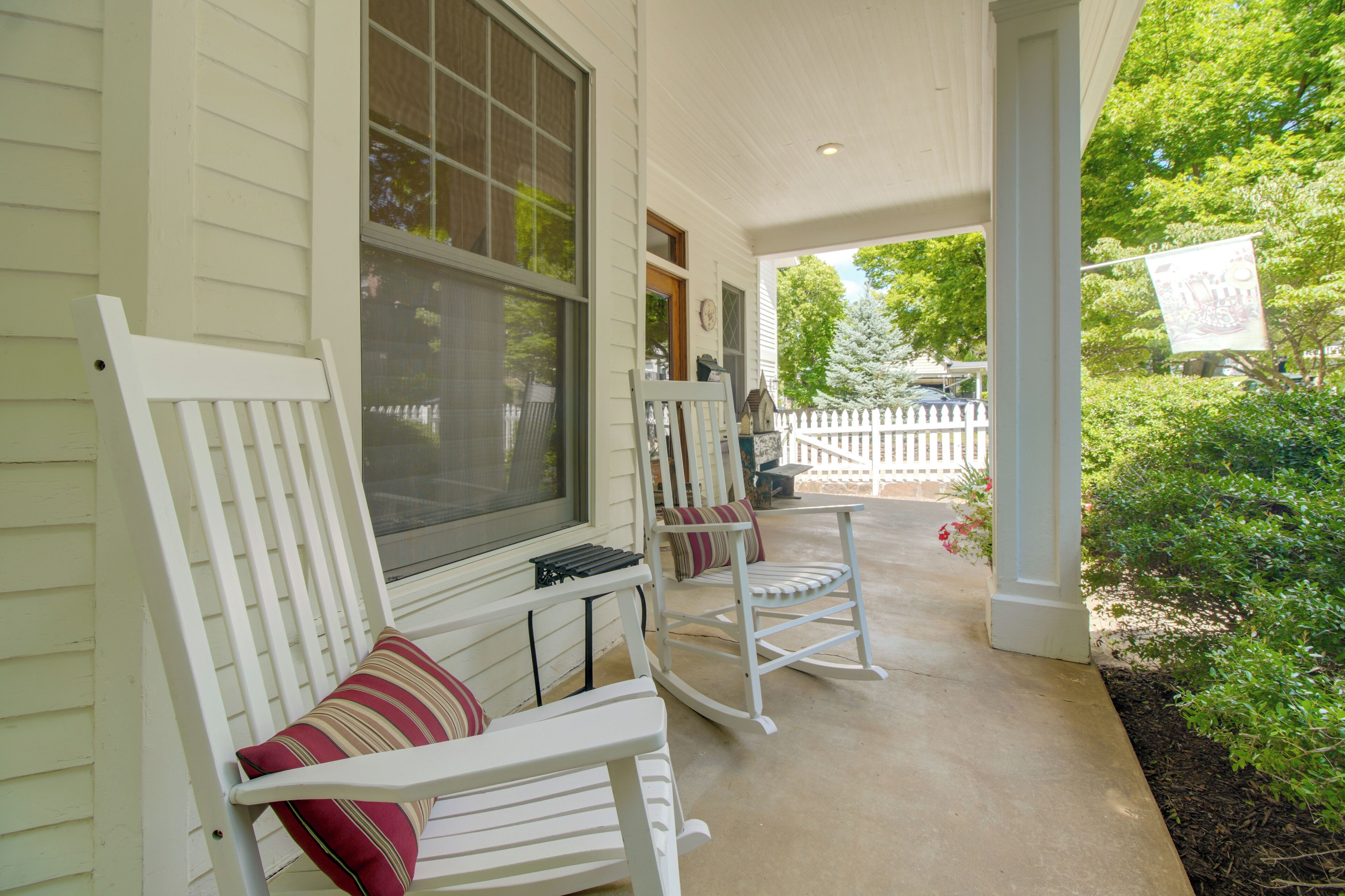 Une véranda avec deux fauteuils à bascule et une petite table, une clôture blanche en bois et un jardin verdoyant.