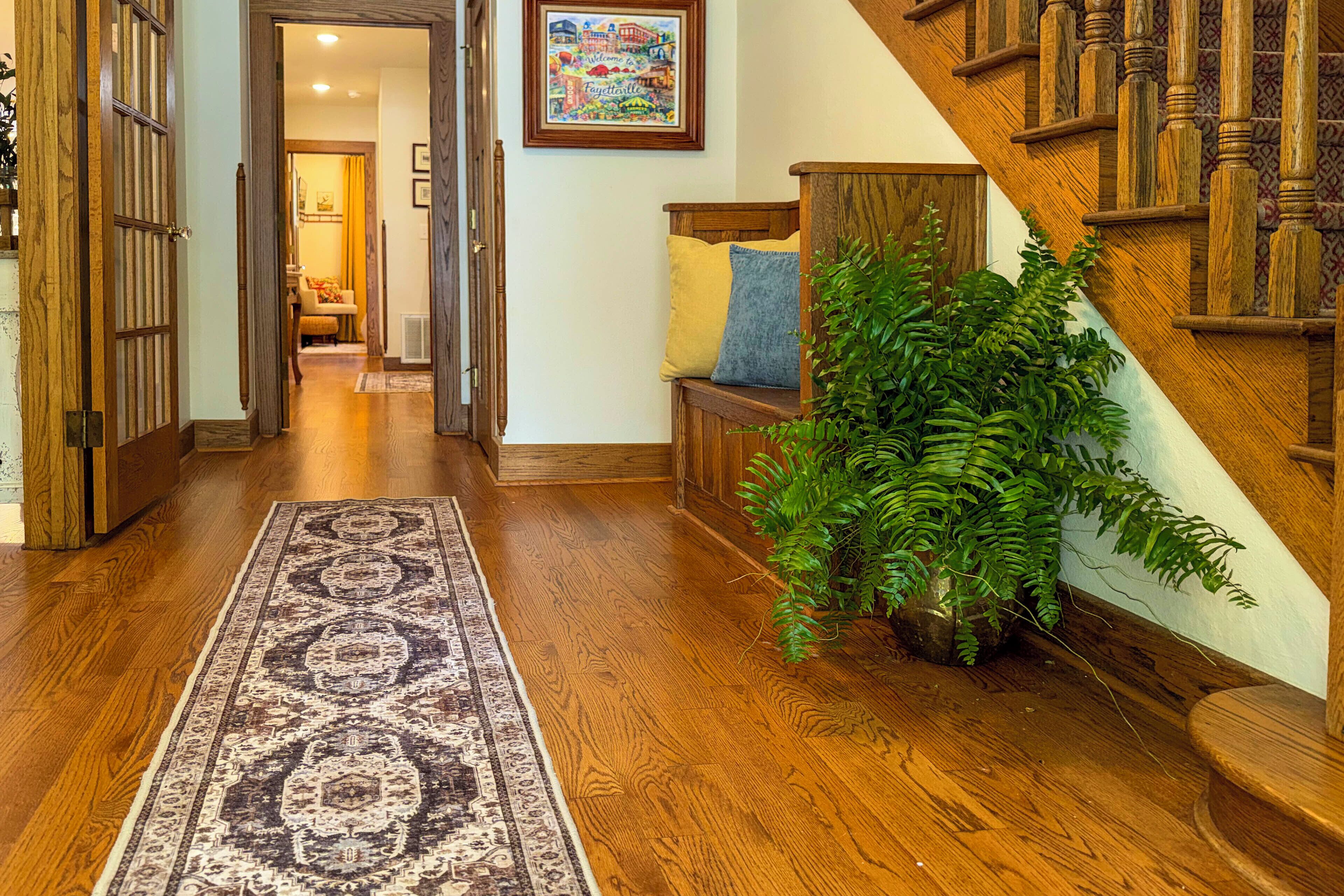 A wooden-floored hallway with a patterned rug, a bench with cushions, and a staircase with wooden railings.