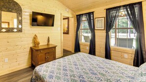 A wooden cabin room with a bed, a dresser, a mirror, a flat-screen TV, and windows with curtains.