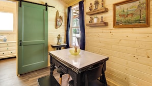 A wooden interior with a table, chairs, a green door, a window with curtains, and wall decorations including figurines and a painting.