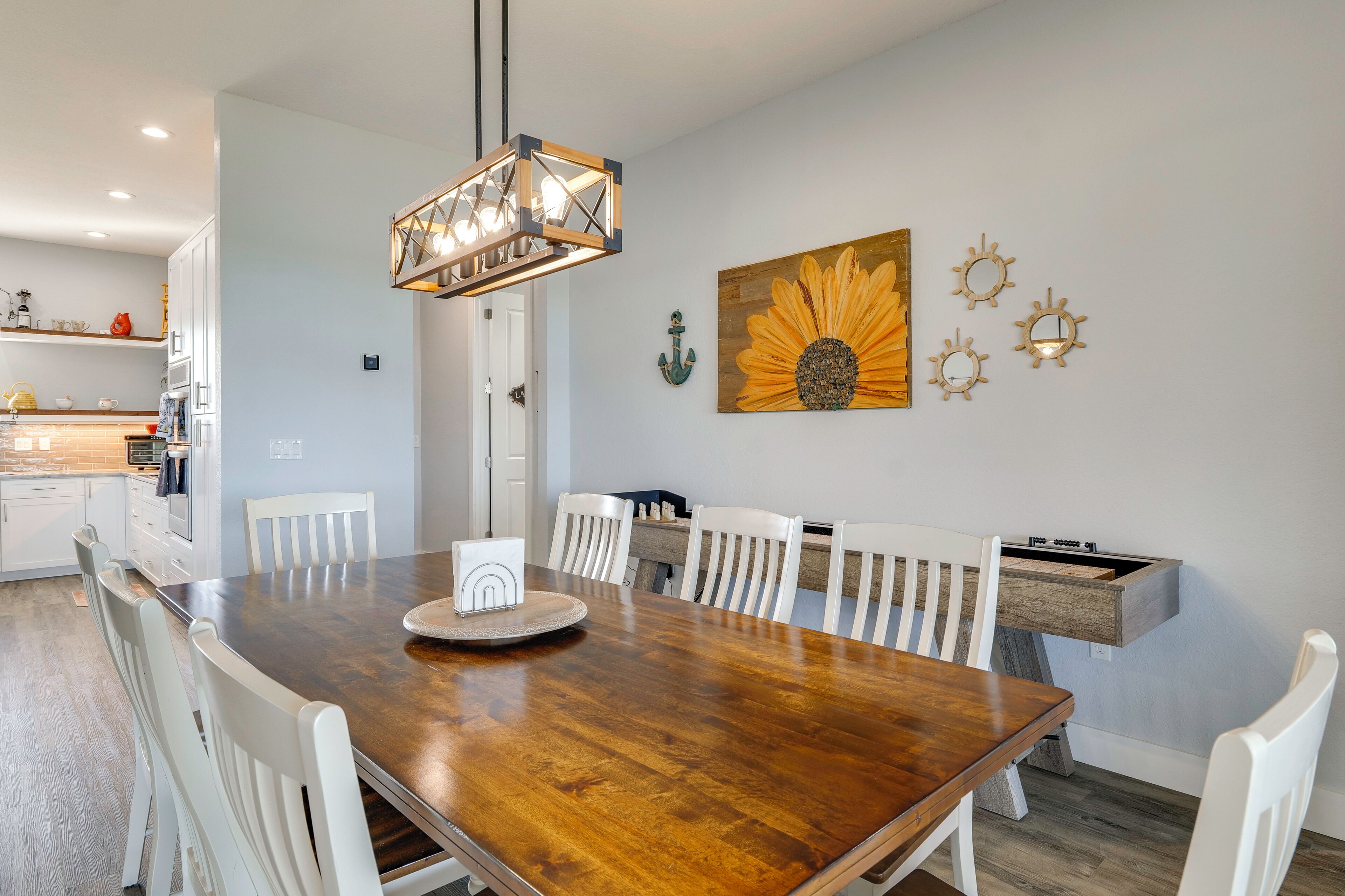 A dining area with a wooden table and white chairs, a pendant light, and wall decor including a sunflower painting and nautical-themed items.