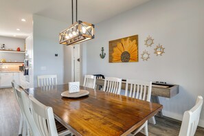 Ein Essbereich mit Holztisch und weißen Stühlen, einer Pendelleuchte sowie Wanddekoration, darunter ein Sonnenblumenbild und maritime Elemente.