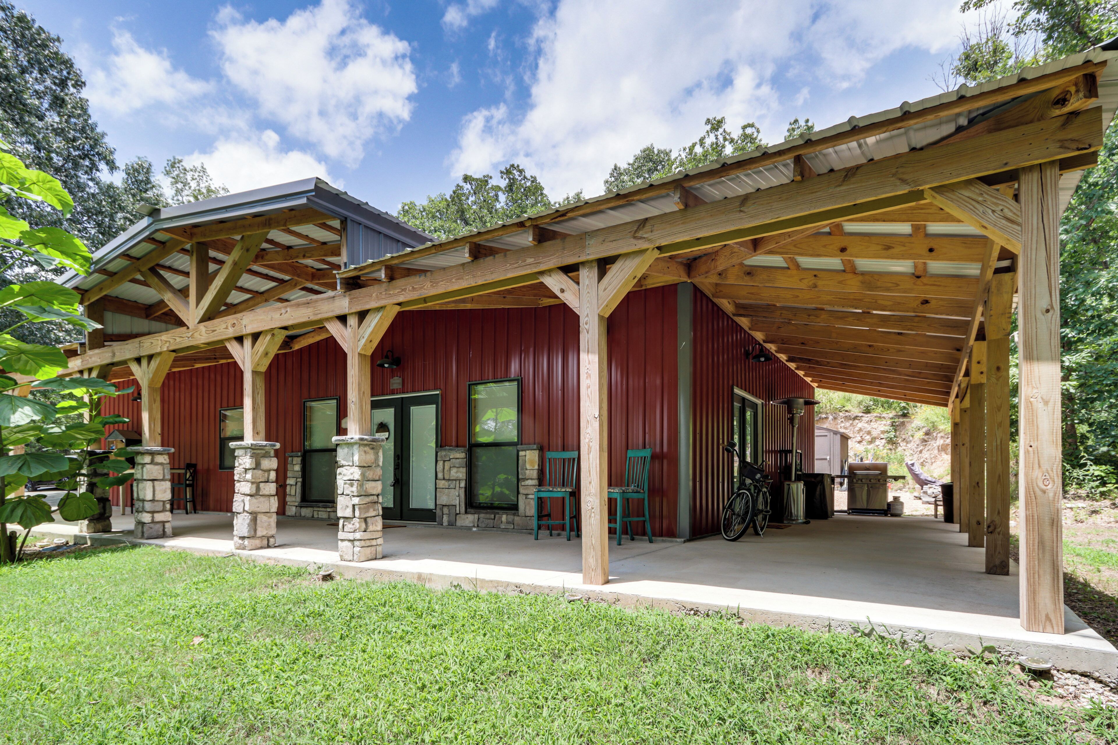 A covered outdoor area with a wooden structure, stone pillars, and a red building in the background.