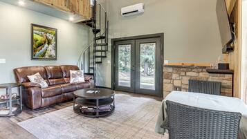 A spacious living area with a leather sofa, a round coffee table, and a wicker chair. There is a staircase leading to an upper level, and a large window with a view of trees.