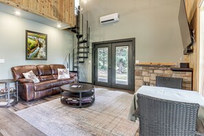 A spacious living area with a leather sofa, a round coffee table, and a wicker chair. There is a staircase leading to an upper level, and a large window with a view of trees.