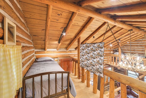 Interior de una cabaña de troncos con una cama, un tapiz colgado y una araña.