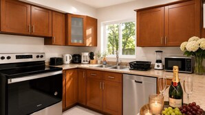 A modern kitchen with wooden cabinets, stainless steel appliances, a large window, and a countertop with a bottle of champagne, grapes, and a candle.