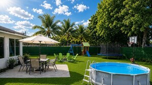 An outdoor swimming pool with a ladder, surrounded by a grassy lawn, a patio with chairs, and a children's play area with a slide.
