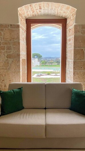Ein Zimmer mit einer Steinwand, einem Fenster mit Blick auf eine Stadtansicht und einem beigefarbenen Sofa mit grünen Kissen.