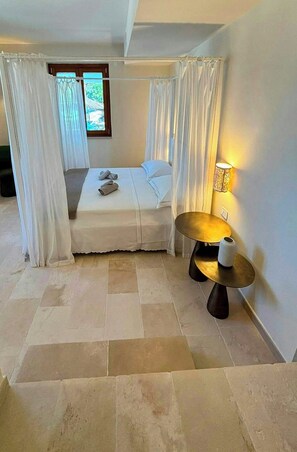 Ein Schlafzimmer mit einem Bett, Vorhängen, einem Nachttisch mit Lampe und Blick auf Grün durch ein Fenster.