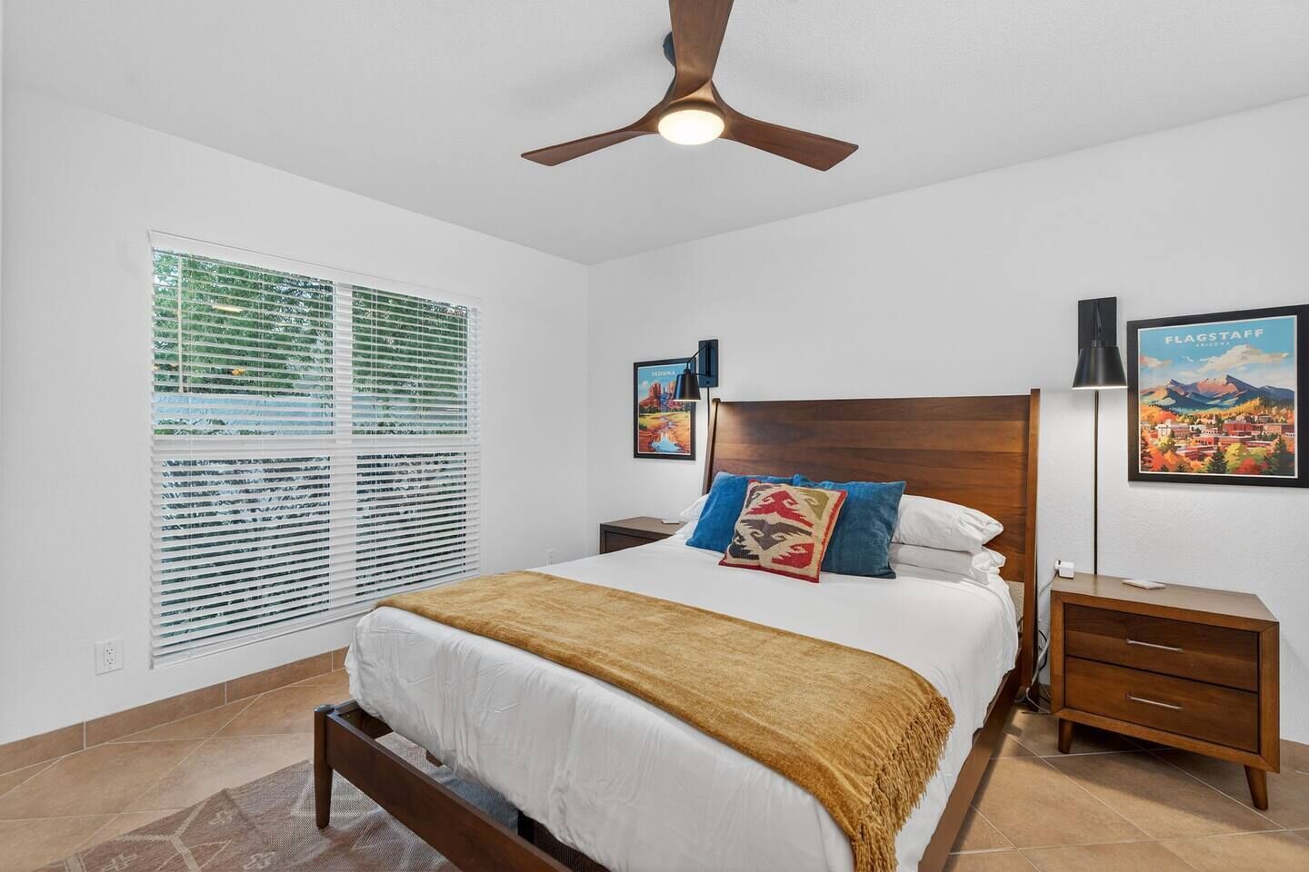 A bedroom with a bed, a ceiling fan, a nightstand, a lamp, and a window with blinds.