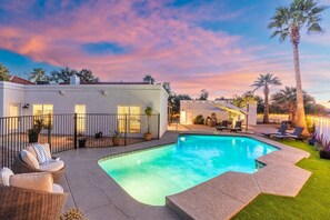 Uma piscina com espreguiçadeiras, um espaço de estar coberto e um edifício branco com janelas.