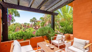 Um pátio ao ar livre com uma mesa de madeira, cadeiras brancas e vista para um jardim exuberante e edifícios.
