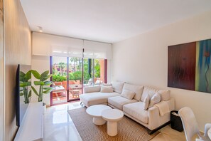 Ein modernes Wohnzimmer mit einem Ecksofa, einem Flachbildfernseher, einem Couchtisch und einem großen Fenster mit Blick ins Freie.