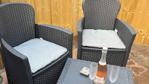 Twee rieten stoelen met grijze kussens, een tafeltje met een fles wijn en twee glaasjes, en een houten hek op de achtergrond.