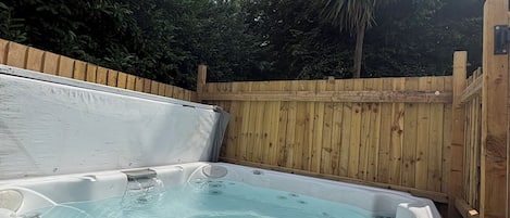 A hot tub with a wooden deck surrounding it, set against a backdrop of trees and a clear sky.