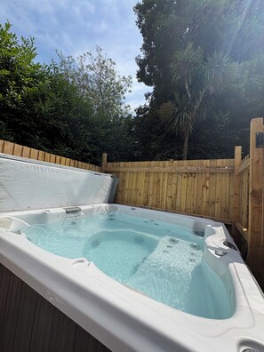 Ein Whirlpool mit umlaufender Holzterrasse, vor einer Kulisse aus Bäumen und blauem Himmel.