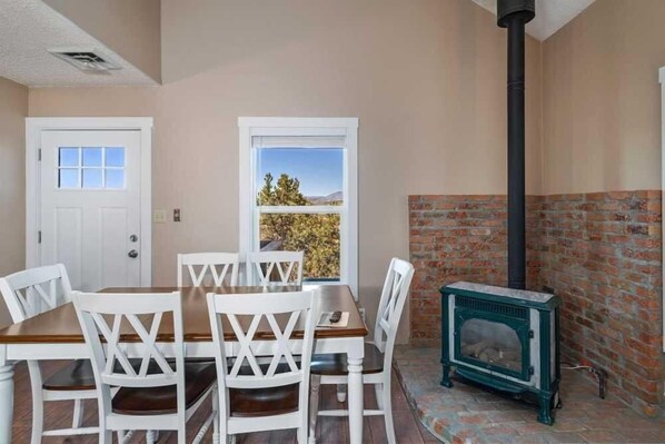 A dining area with a brick fireplace, white chairs, a wooden table, and a view through a window.