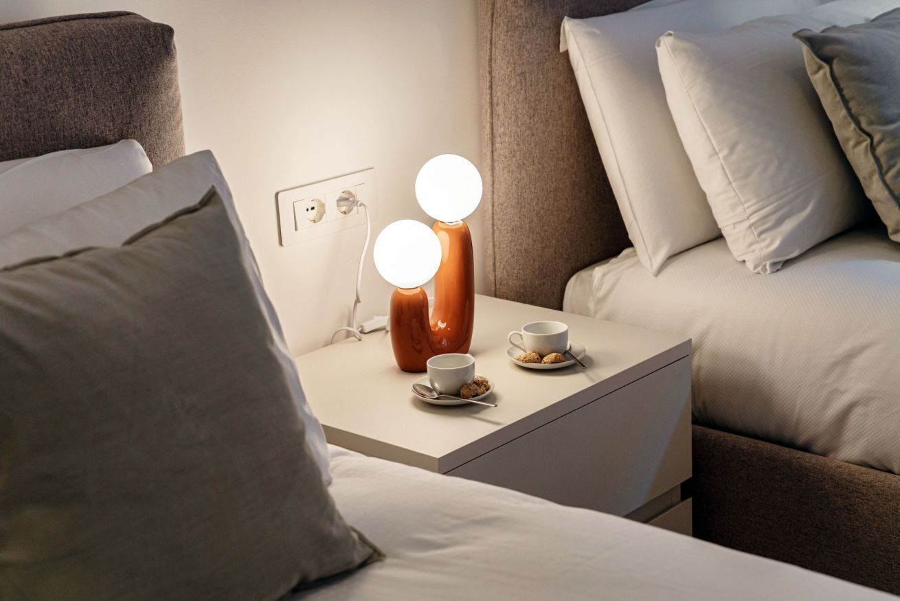 Ein ordentlich bezogenes Bett mit Kissen, ein Nachttisch mit Lampe und eine Tasse Kaffee darauf.