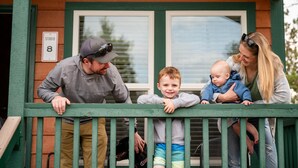 Una familia en un porche frente a una casa de madera, una ventana y un letrero que dice 'STUDIO 8'.