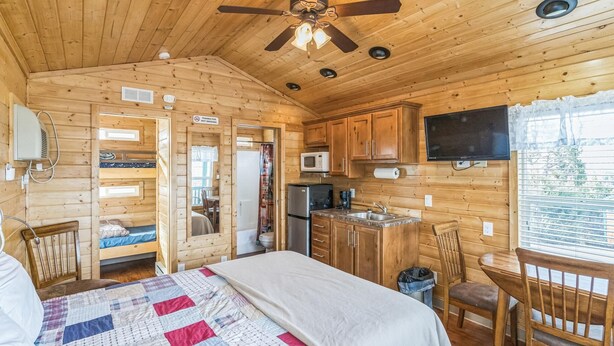 Interior de una cabaña de madera con cama, kitchenette y ventilador de techo.