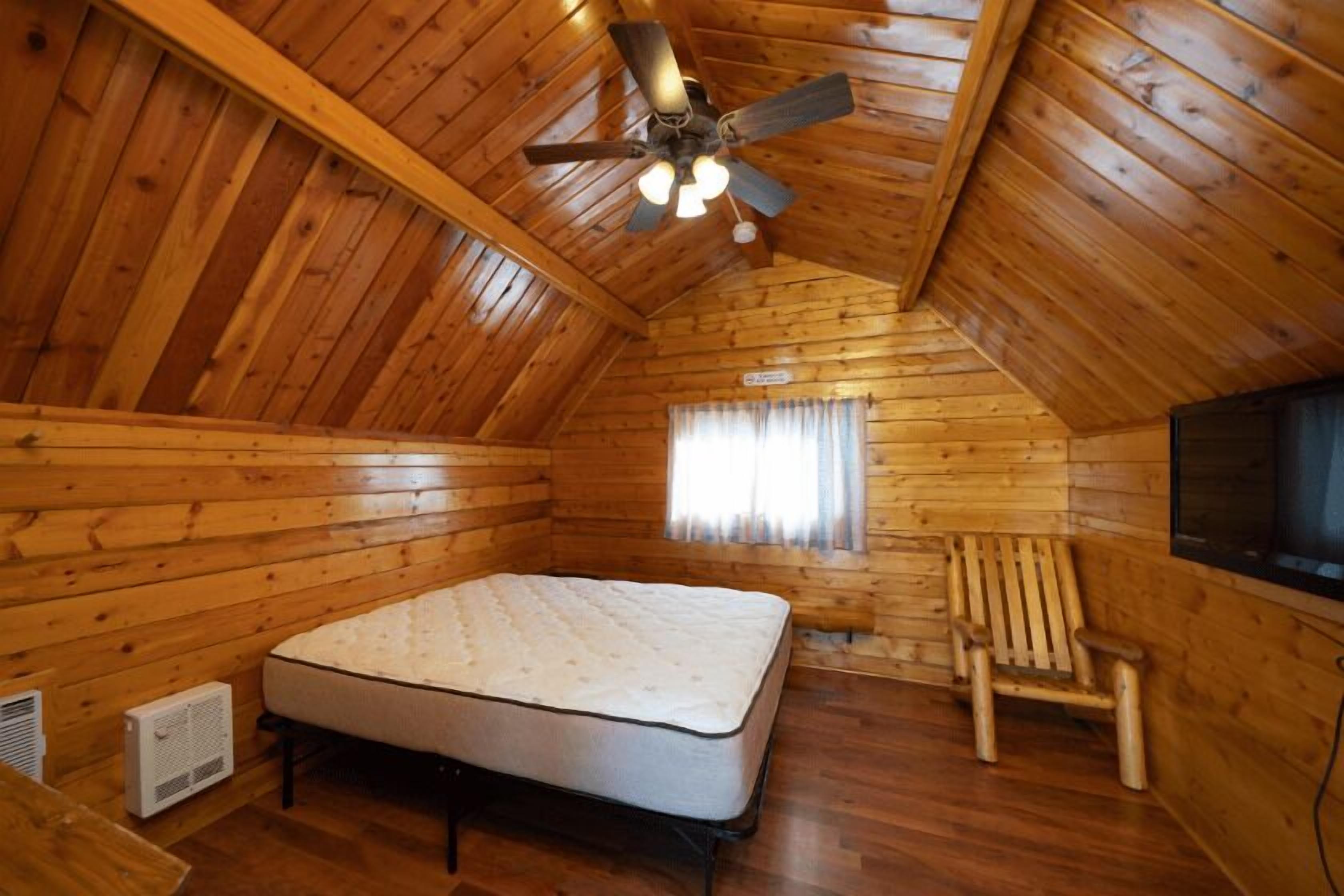 Una habitación de cabaña de madera con una cama, ventilador de techo, calefactor, ventana con cortinas y televisor.
