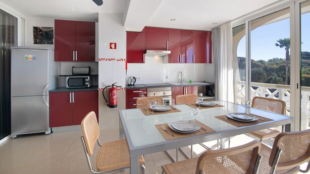 Une cuisine moderne avec des meubles de rangement rouges, une table à manger en verre, des chaises en osier et une vue sur l’extérieur.