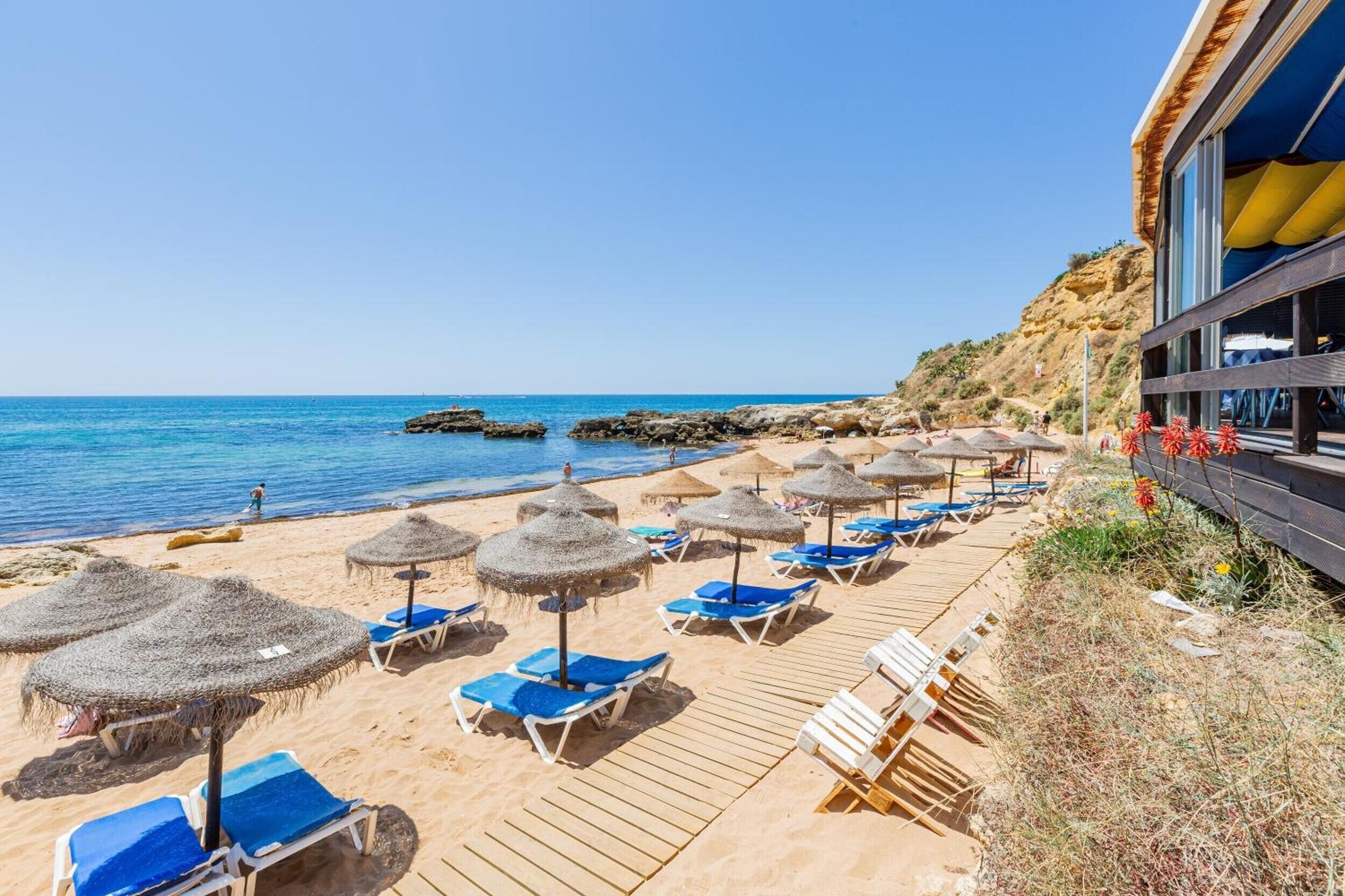 Un hôtel situé en bord de mer, doté d’une terrasse en bois, de chaises longues bleues et de parasols en chaume.