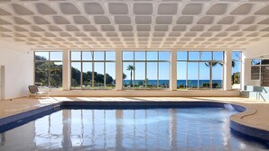 Une grande piscine intérieure avec des bords carrelés, de grandes fenêtres offrant une vue sur la mer et un plafond orné d’un motif géométrique.
