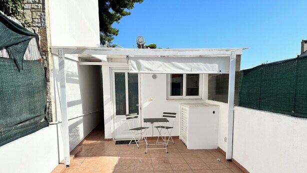 Een klein terras met een witte tafel en stoelen, een compacte koelkast, en een raam met uitzicht op een tuin.