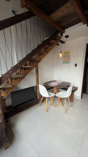 A dining area with a wooden staircase, white chairs, and a table set for two. - Vila Artesania (Bombinhas)
