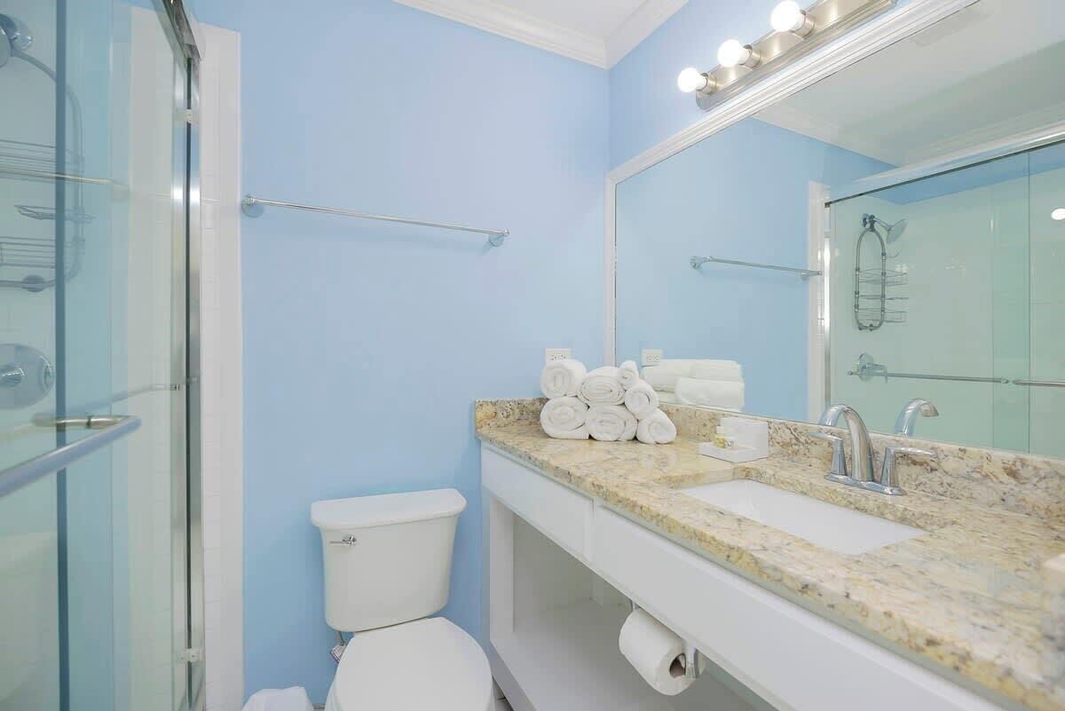 A bathroom with a glass shower enclosure, a toilet, a sink with a marble countertop, and a mirror with lights.