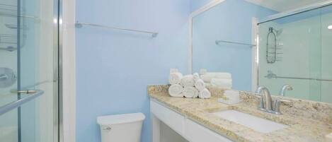 A bathroom with a glass shower enclosure, a toilet, a sink with a marble countertop, and a mirror with lights.