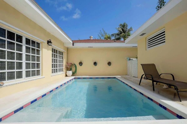 Una piscina con borde de azulejos, una ventana con persianas y una silla.