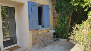 Un edificio con puerta de vidrio corrediza, una pequeña zona de estar exterior con mesa y sillas, y una ventana con contraventanas azules.