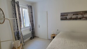 Un dormitorio con una cama, una ventana con cortinas, una puerta, un cuadro en la pared y una mesita de noche con una vela.