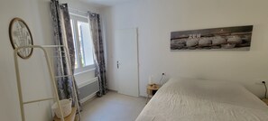 Ein Schlafzimmer mit einem Bett, einem Fenster mit Vorhängen, einer Tür, einem Wandbild und einem Nachttisch mit einer Kerze.