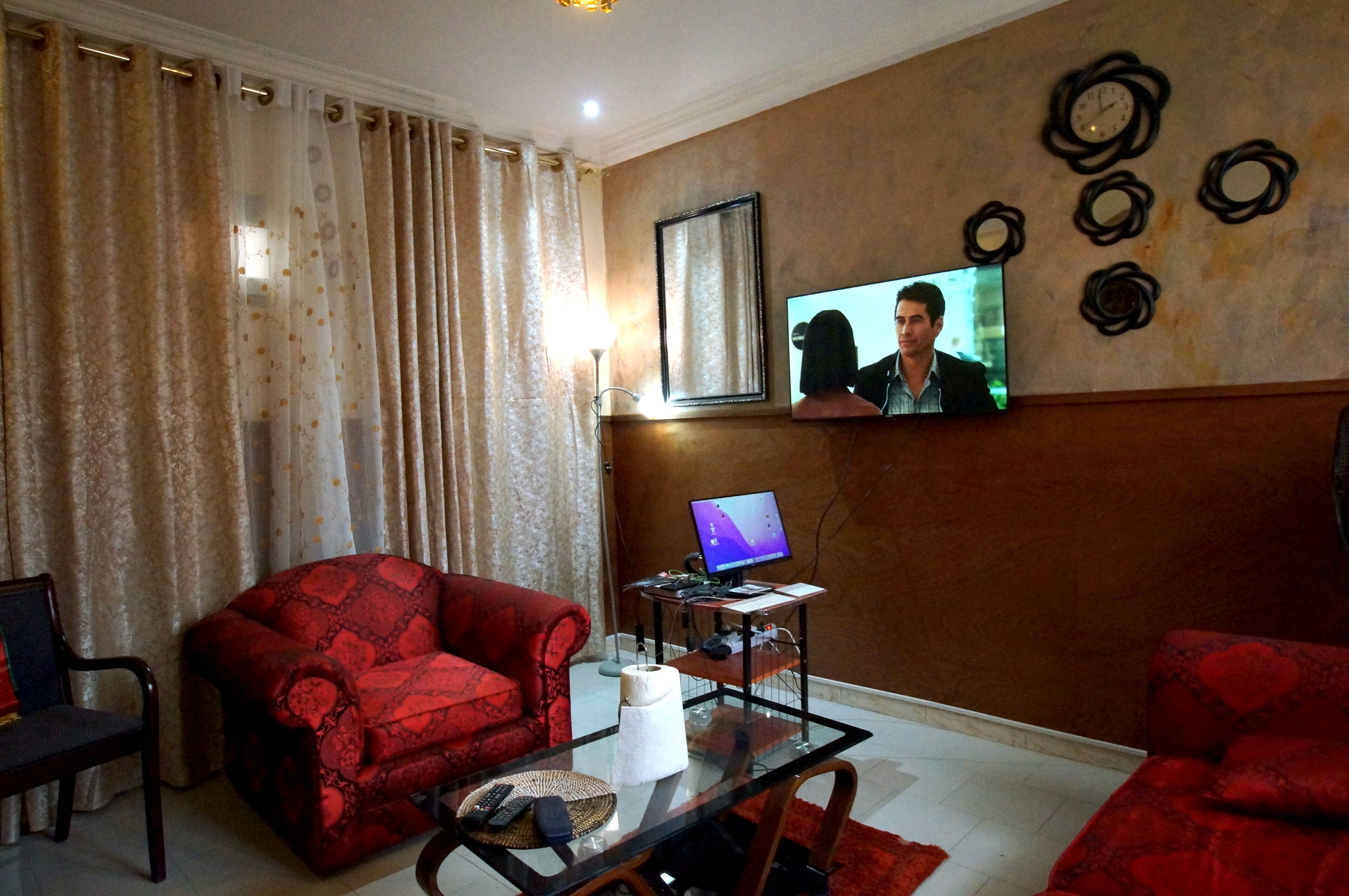 A living room with a red sofa, a glass coffee table, a TV mounted on the wall, and a computer on a small desk.