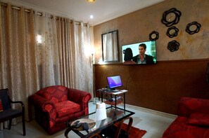 Ein Wohnzimmer mit einem roten Sofa, einem Glastisch, einem an der Wand befestigten Fernseher und einem Computer auf einem kleinen Schreibtisch.