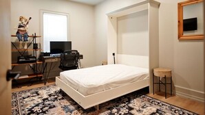 Une chambre à coucher comprenant un lit, un bureau, une chaise, un petit tabouret, un ventilateur de plafond et une fenêtre.